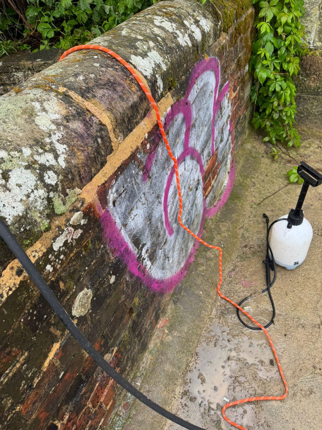 Graffiti on a brick wall before removal, with pressure-washing equipment ready, in West Sussex